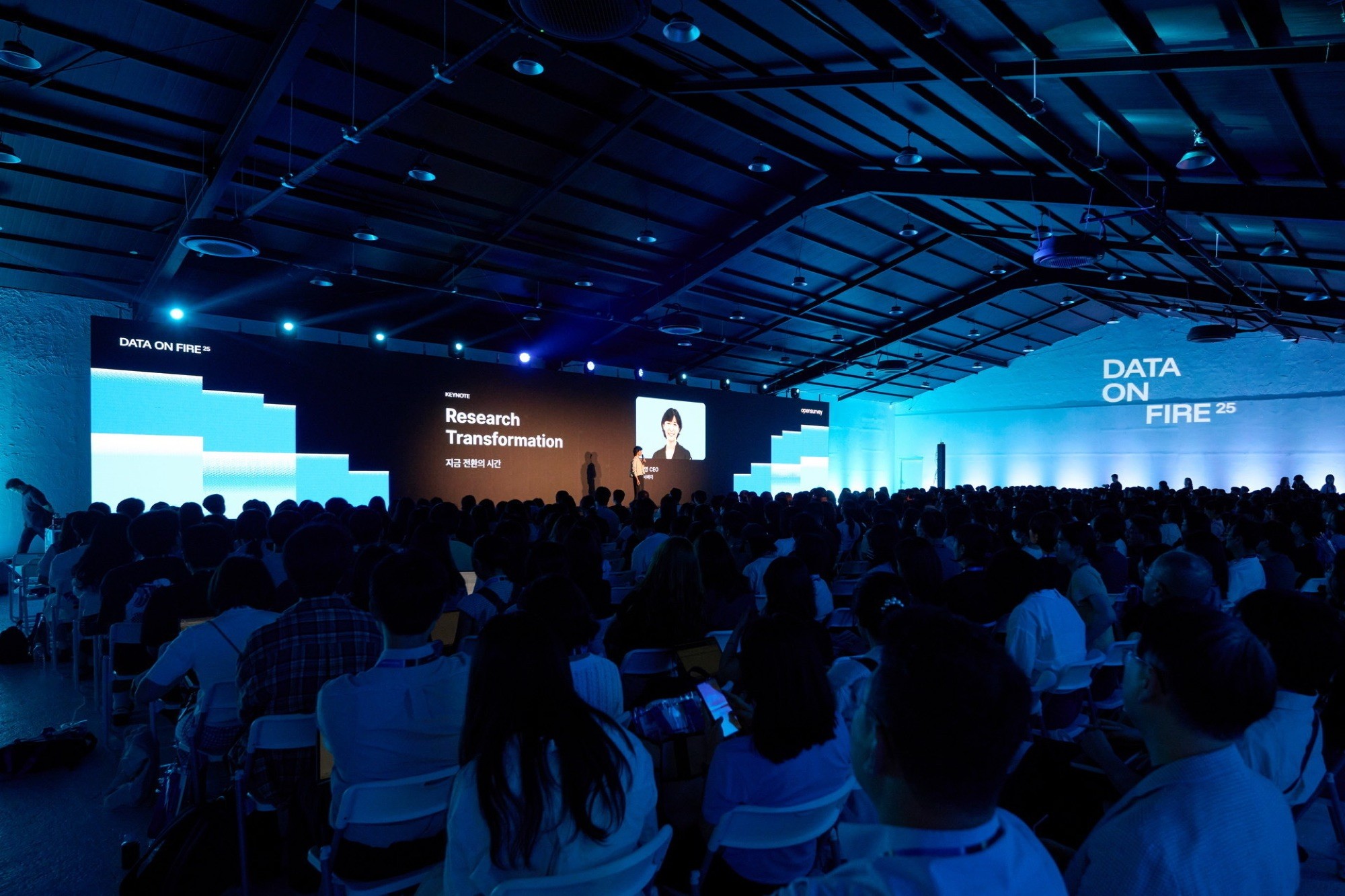 Audience gathered in a dimly lit event space, with blue lighting and a large screen in the foreground.