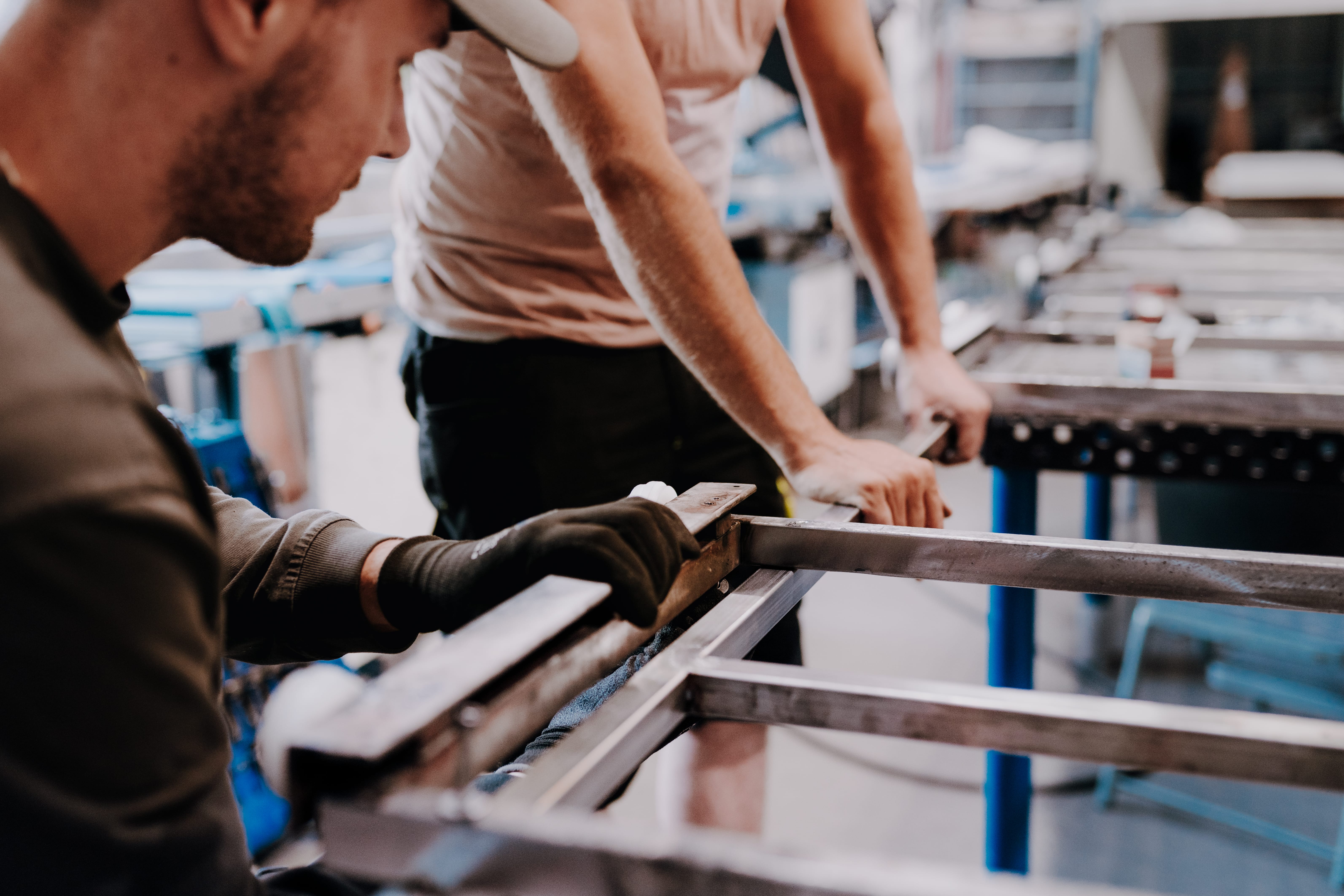 Two workers are assembling metal frames in a workshop. One is working on a frame with gloved hands.