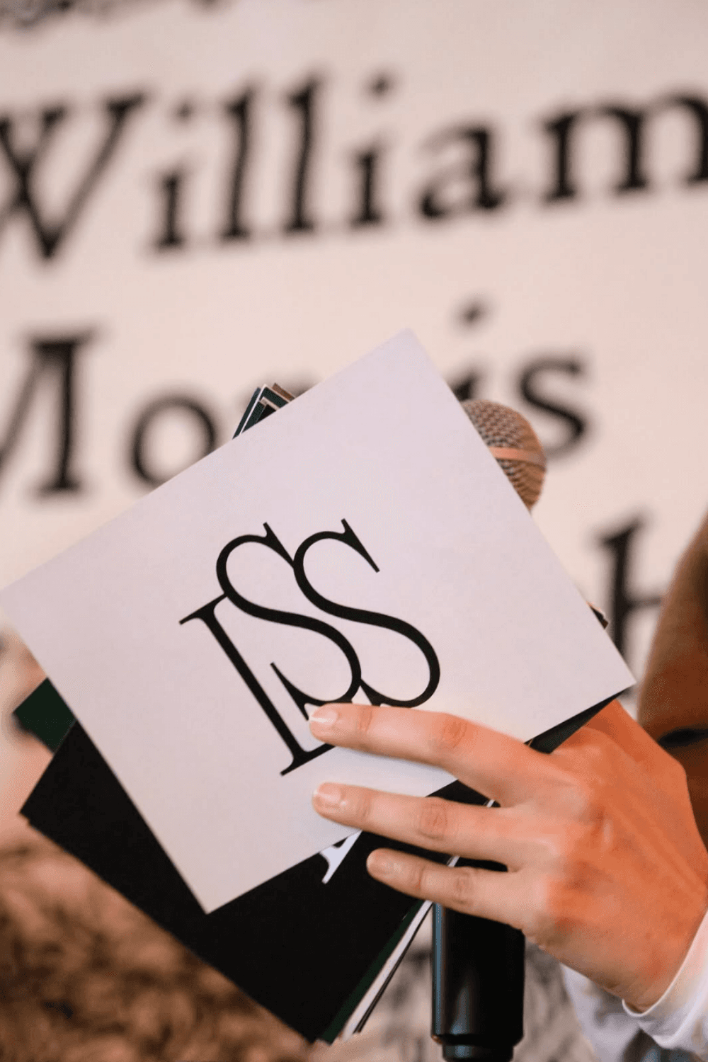 A person holding playing cards with a contemplative expression, smoking a cigarette. A glass and ashtray are on the dimly lit table, creating a tense mood.