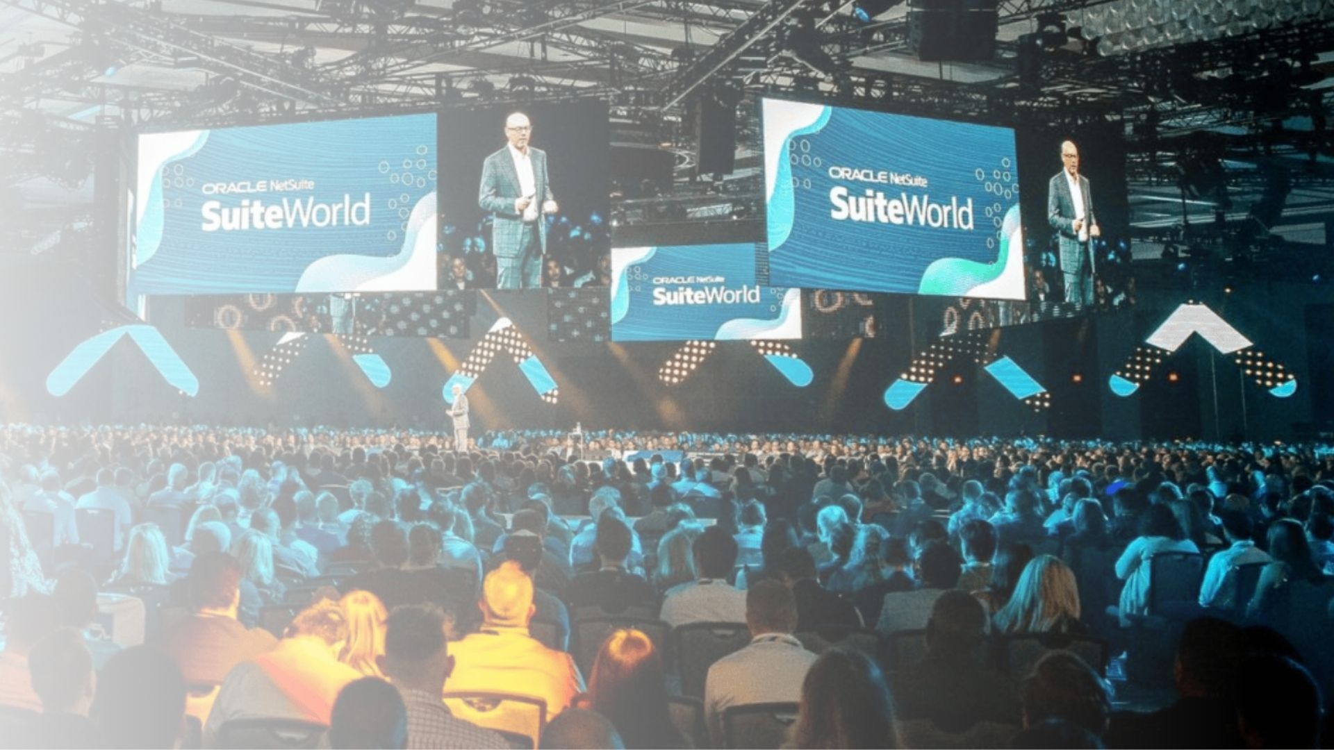 Audience sitting in a conference hall with large screens displaying text, illuminated by stage lighting.
