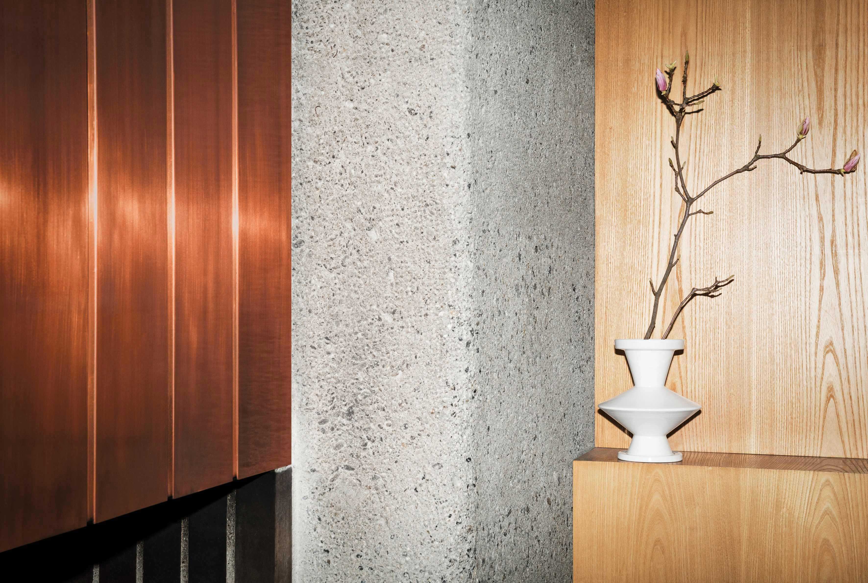  A close-up shot of architectural details. On the left are a series of vertical copper panels. In the center is a textured concrete column. On the right, a simple white vase with a flowering branch is placed on a wooden shelf against a wooden wall.