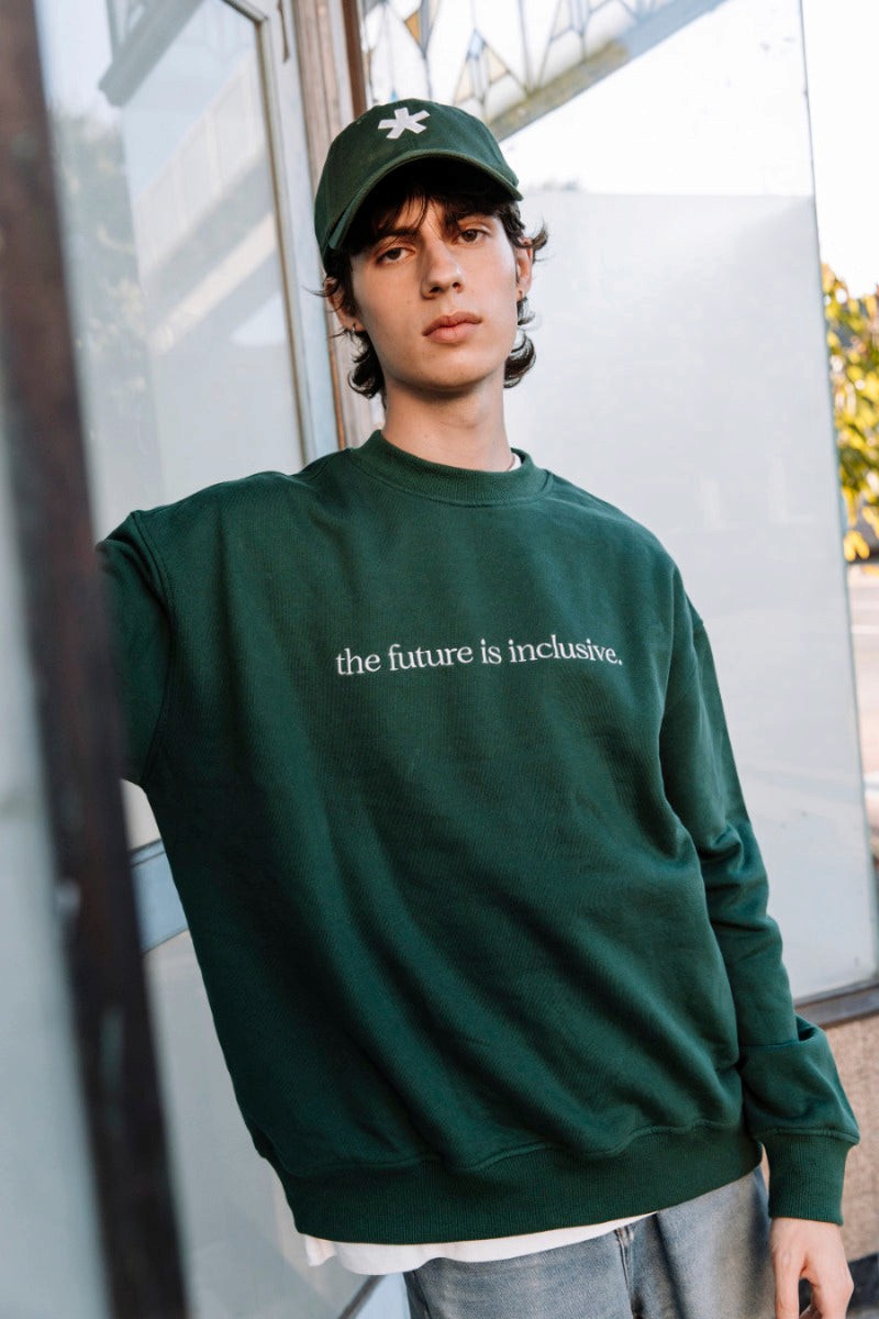 Person wearing a hunter green sweatshirt with 'the future is inclusive' text, standing outdoors.