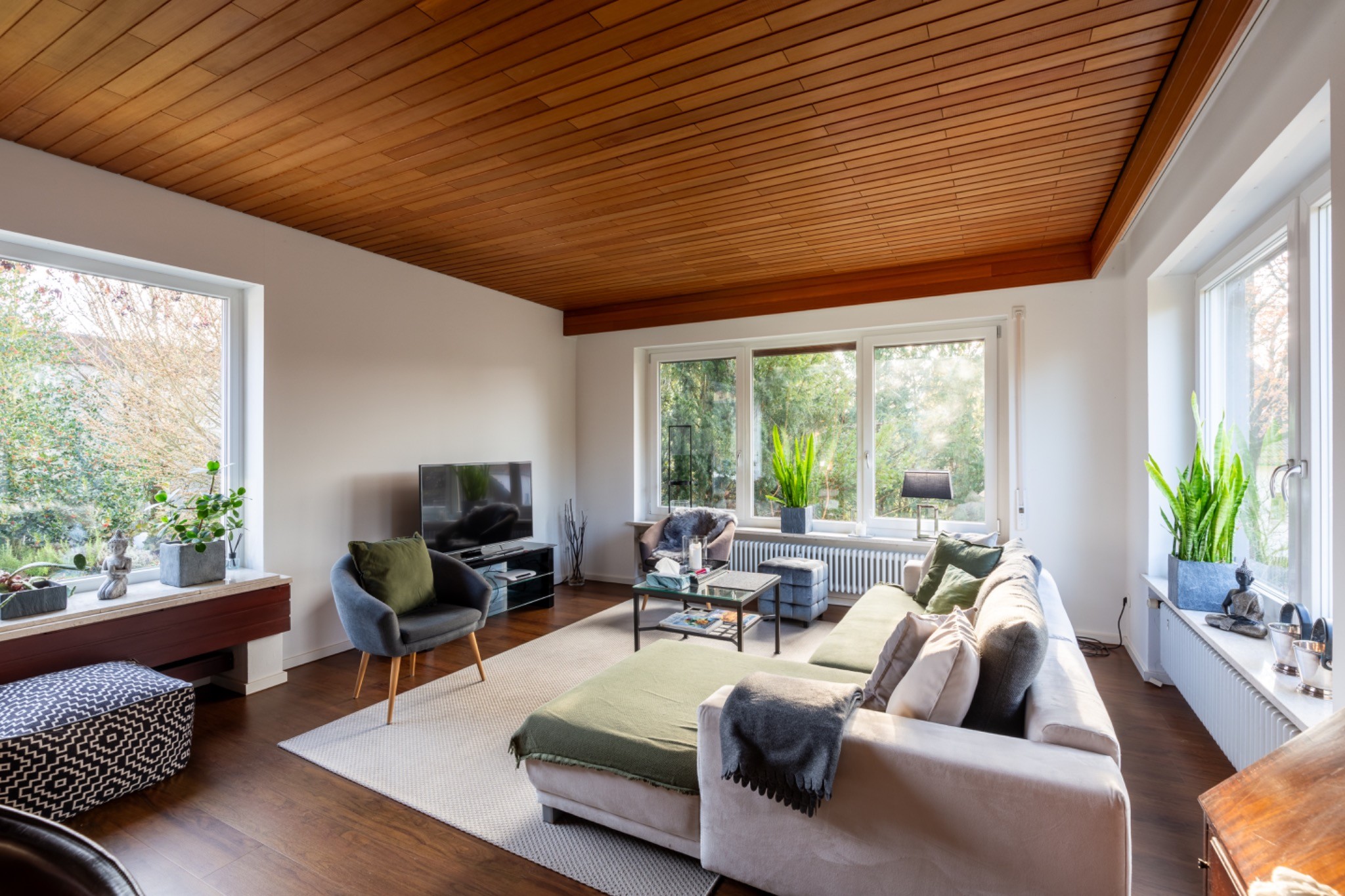 Wohnzimmer mit Holzdecke, großen Fenstern auf mehreren Seiten, einer Sofaecke und Blick ins Grün.