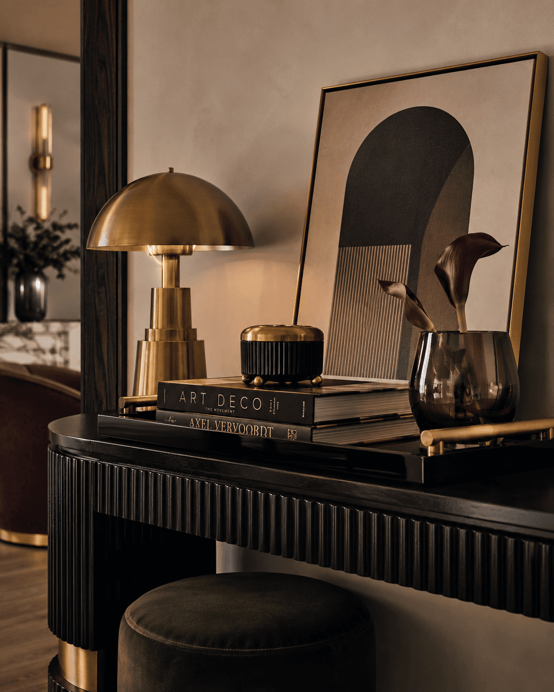 Close-up Neo Deco interior vignette featuring a black fluted console table styled with a sculptural brass lamp, glossy lacquer tray, smoked glass vase, stacked Art Deco design books, and an arched framed artwork. Rich material layering includes polished brass, velvet, walnut wood, marble, and lacquer finishes. Warm ambient lighting creates soft reflections and a moody luxury atmosphere inspired by 2026 Art Deco revival interior design trends.
