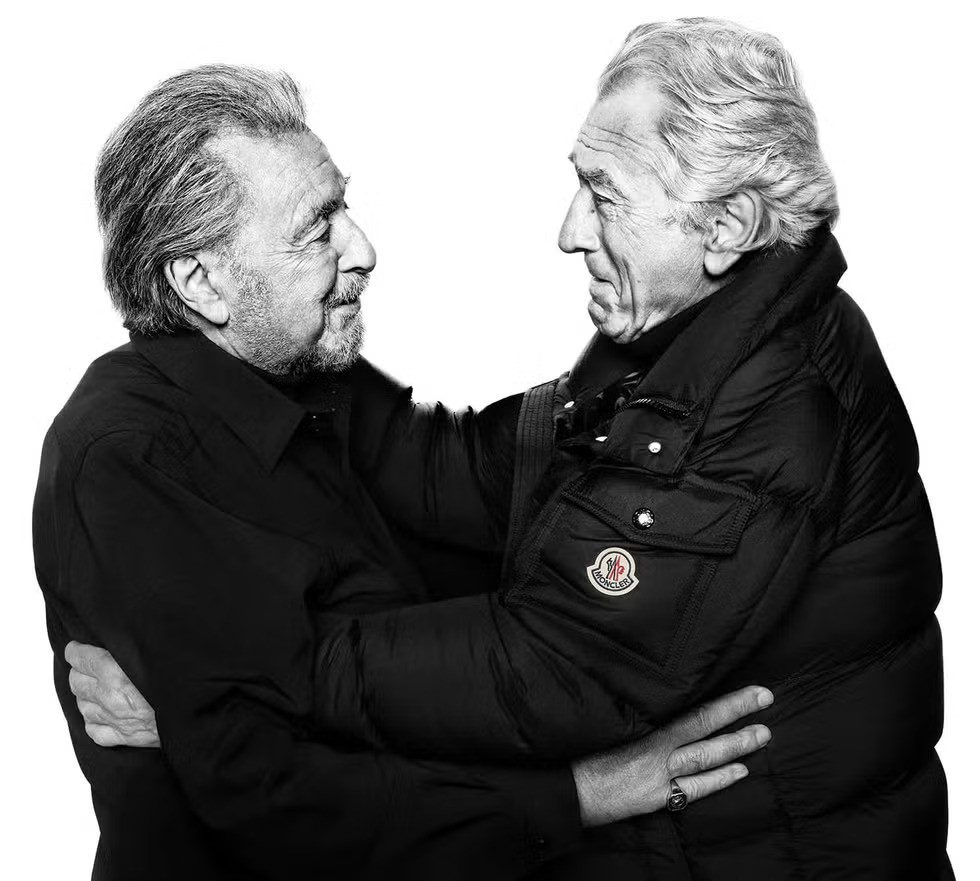 Robert De Niro and Al Pacino embracing each other while seen black Moncler outerwear, photographed in a minimalist black-and-white studio setting.
