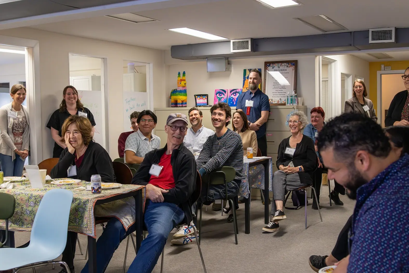 A group of people sitting and standing in a brightly lit room, appearing engaged and smiling, with tables and chairs arranged for a social gathering, suggesting a casual meeting or community event.