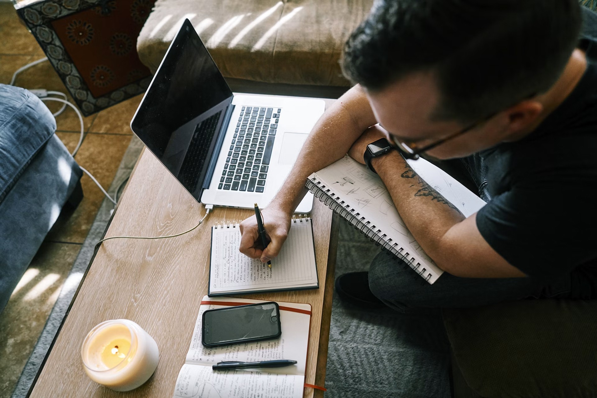 Person writing in a notebook beside a laptop and smartphone, showing focused productivity with minimalist phone.
