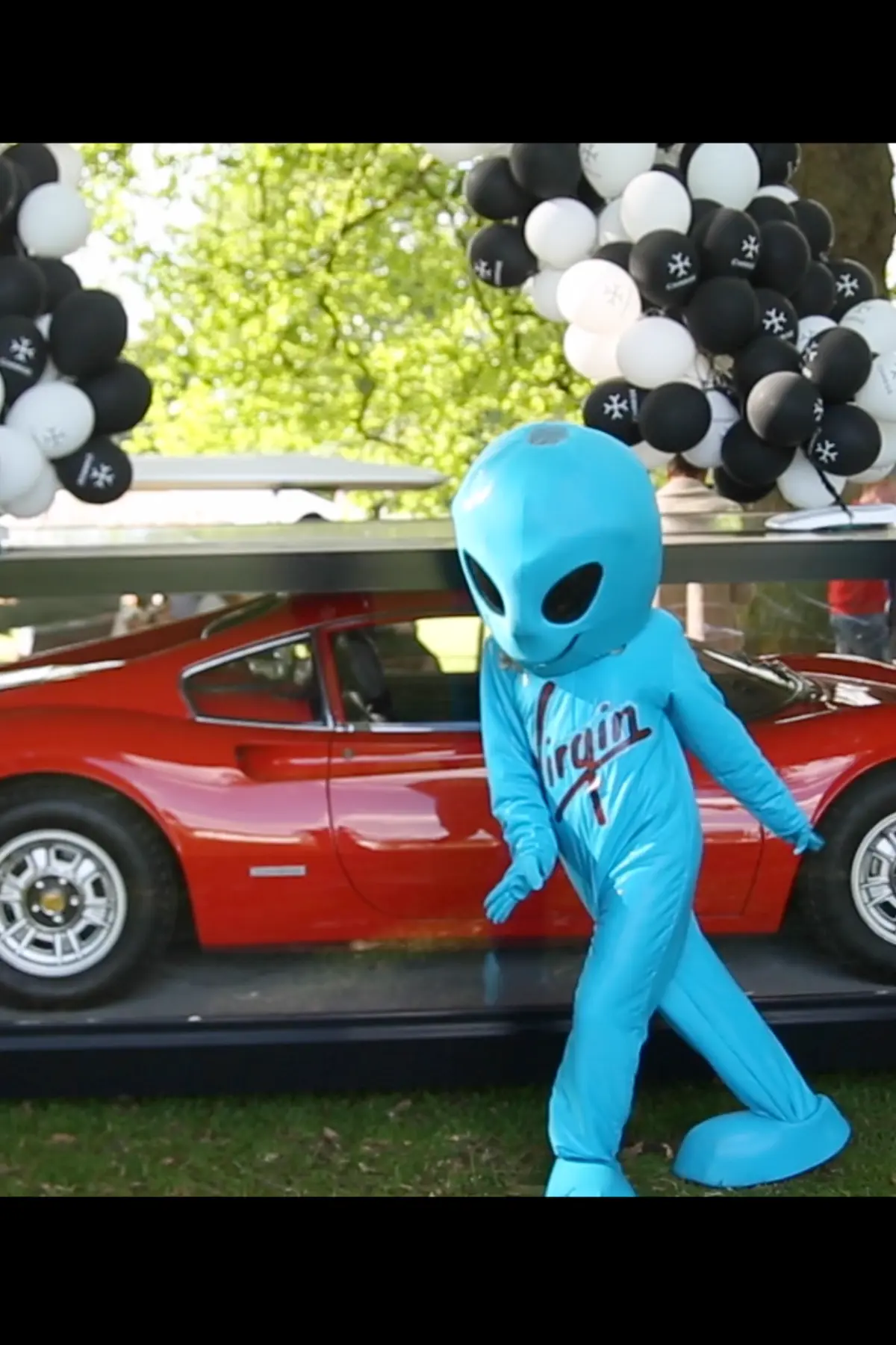 Agent Blonde dancing in a Virgin alien costume in front of a red car decorated with balloons