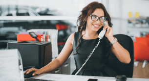 A smiling woman with glasses on the phone at a desk with a computer.
