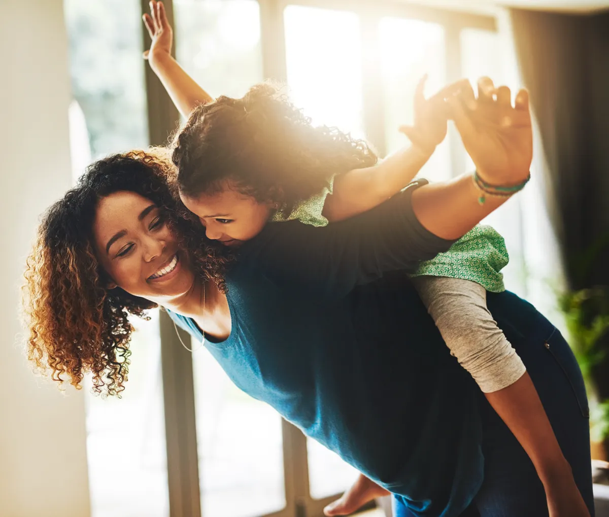 Happy mother giving her young daughter a piggyback ride indoors with sunlight streaming through the windows.