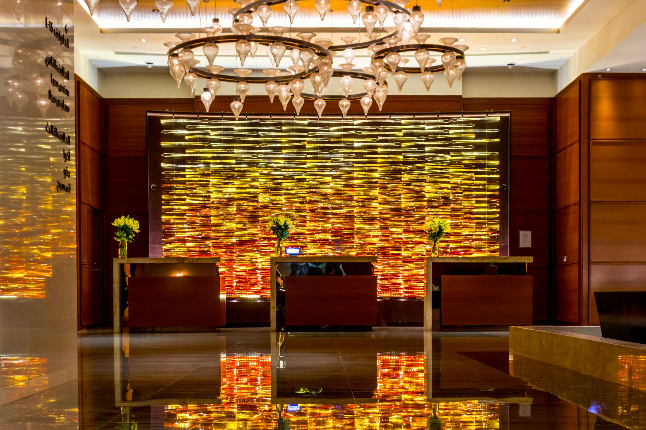 A modern hotel lobby features three reception desks in front of a textured gold and orange backlit wall under a crystal