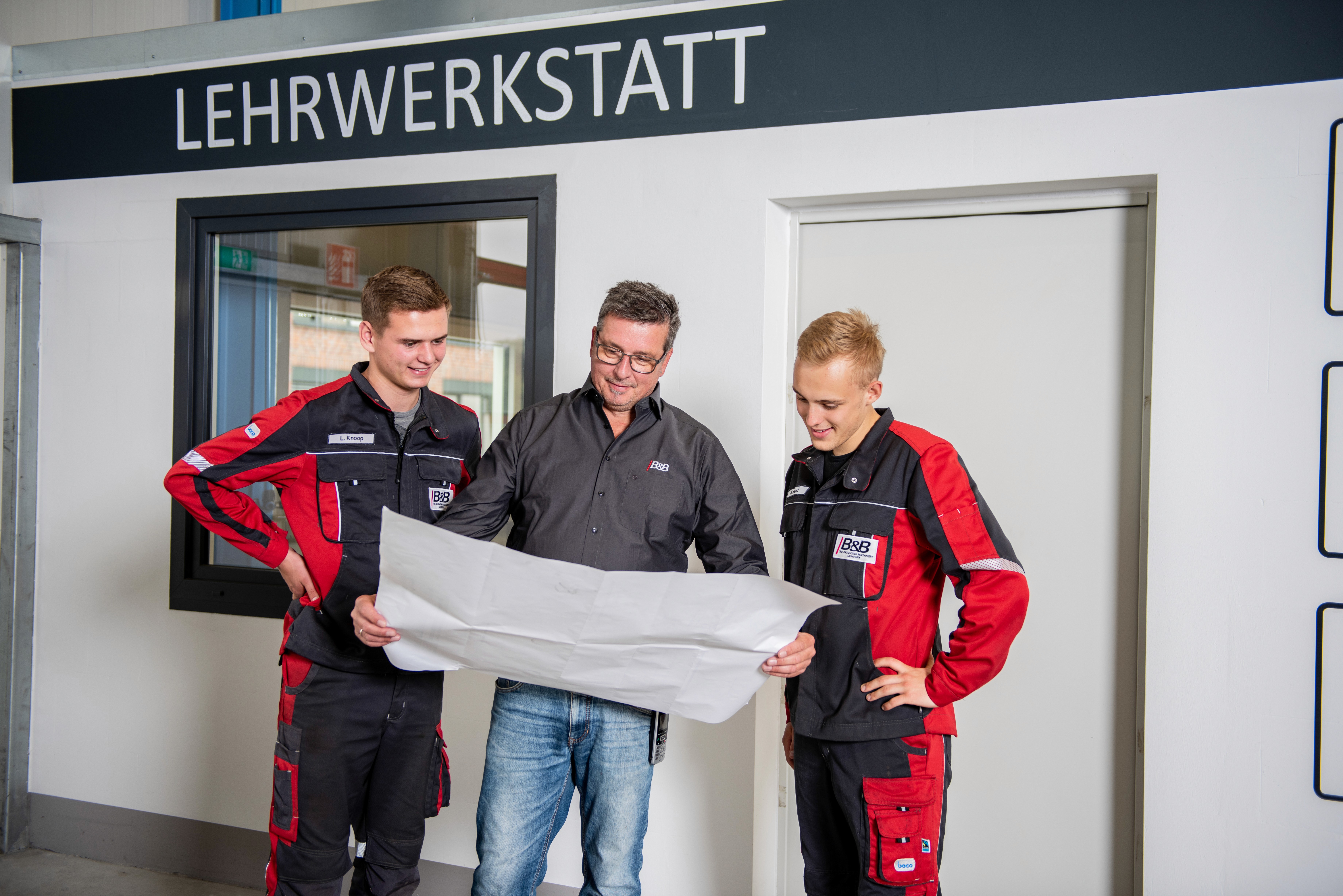 Lehrer mit Auszubildenden in der Lehrwerkstatt, Bauzeichnung prüfend. Berufsausbildung.