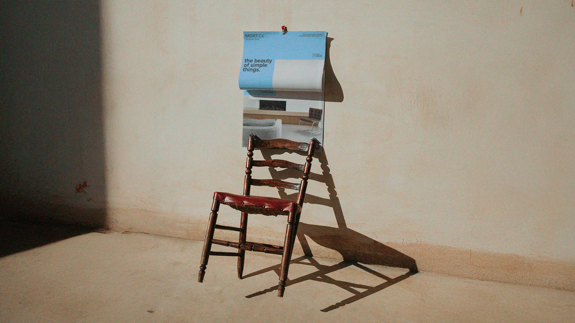 Une scène intérieure minimaliste, baignée de lumière naturelle, où une vieille chaise en bois projette une ombre allongée sur un mur texturé. Au-dessus, une affiche aux tons bleus porte l’inscription « the beauty of simple things ». La lumière chaude et le vide volontaire de l’espace installent une atmosphère calme, contemplative, presque poétique.
