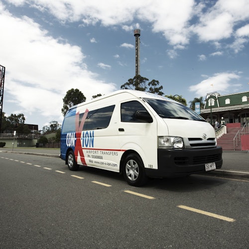 Witte Conn-X-ion luchthaven transferbus geparkeerd op een straat met een groen gebouw en bomen op de achtergrond.