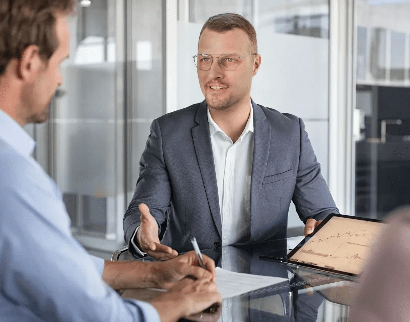 Zwei Geschäftspartner in einem Besprechungsraum; ein Mann mit Brille im Anzug führt ein Gespräch, während sein Gegenüber sich Notizen macht und ein Tablet auf dem Tisch liegt.