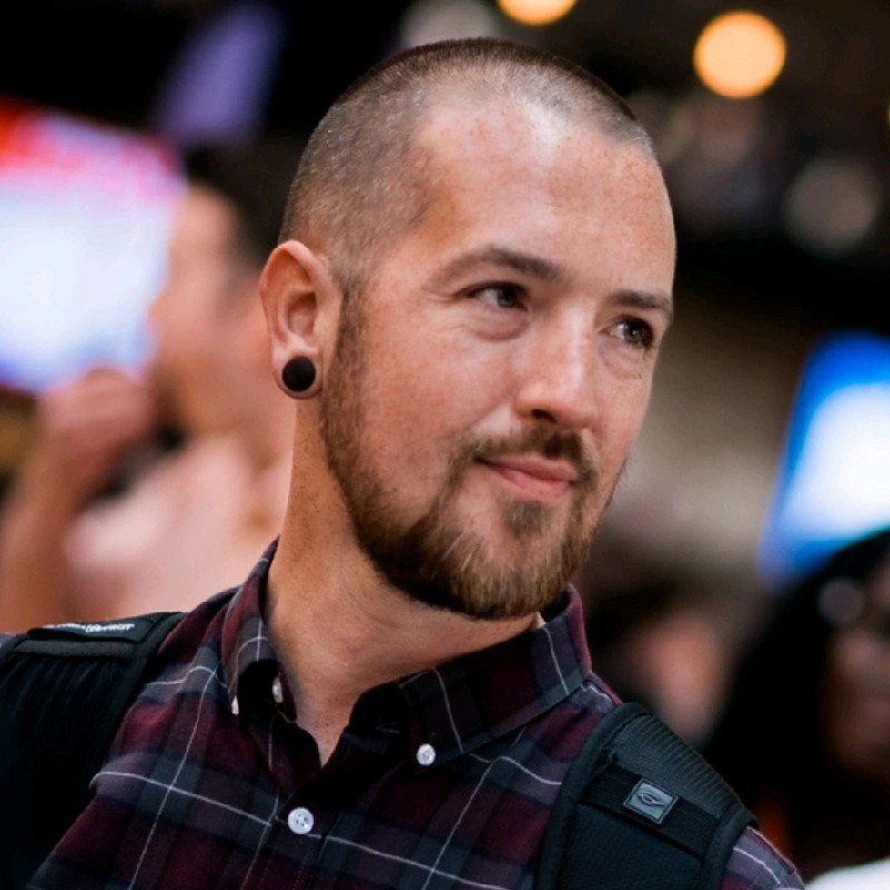 Portrait photograph of a person with closely shaved hair and a short beard, wearing a dark checked shirt and black backpack straps, with a softly blurred indoor background.