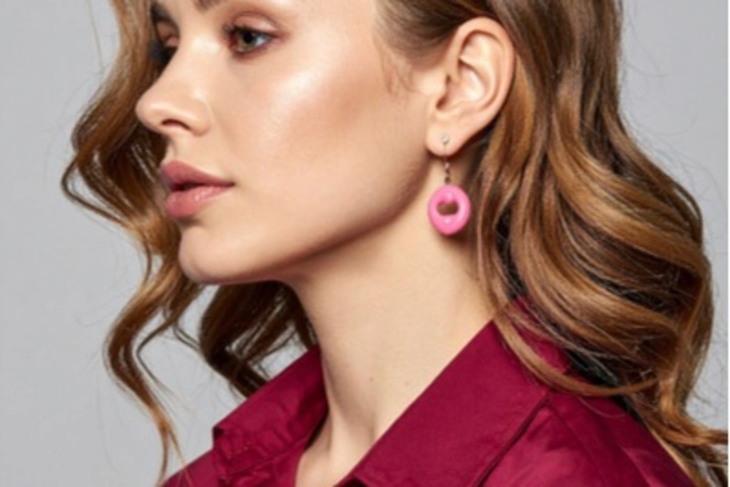 ChatGPT said: A stylish woman with wavy light brown hair wearing a deep burgundy satin blouse and pink circular drop earrings, showcasing a modern, polished look perfect for contemporary fashion inspiration.