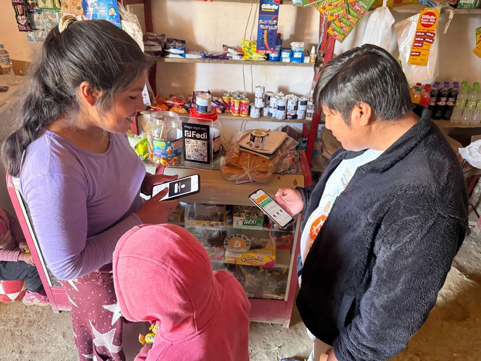 A shopper and a small shop owner in rural Peru complete a Bitcoin payment using Fedi on their phones. A "Paga aquí con Fedi" QR code sign and an "Aceptamos Bitcoin" sticker are visible on the shop counter, stocked with everyday goods.