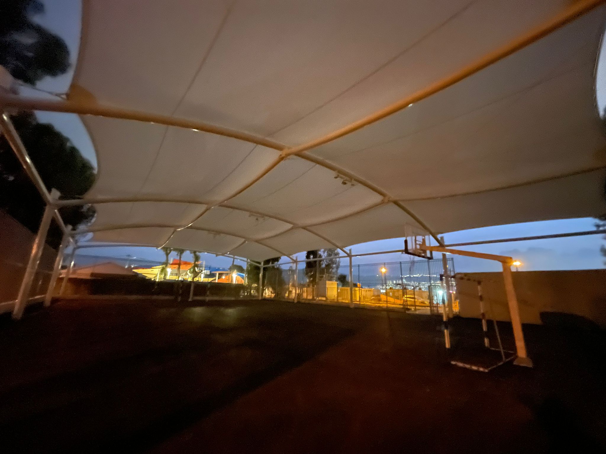 Covered area with beige fabric roof and metal frame at dusk with illuminated buildings visible in the background.