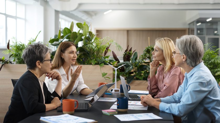 Four adults in a modern office setting engage in a collaborative discussion around a table with laptops and documents, surrounded by lush green plants.