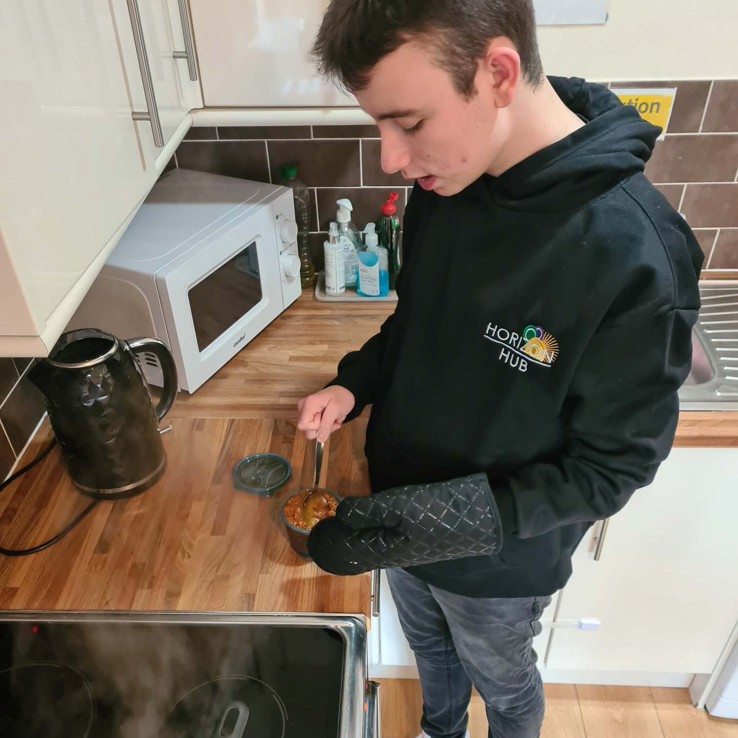 A teenager is stood at a kitchen counter stirring a hot bowl of soup with a sppon. They are wearing an oven glove.