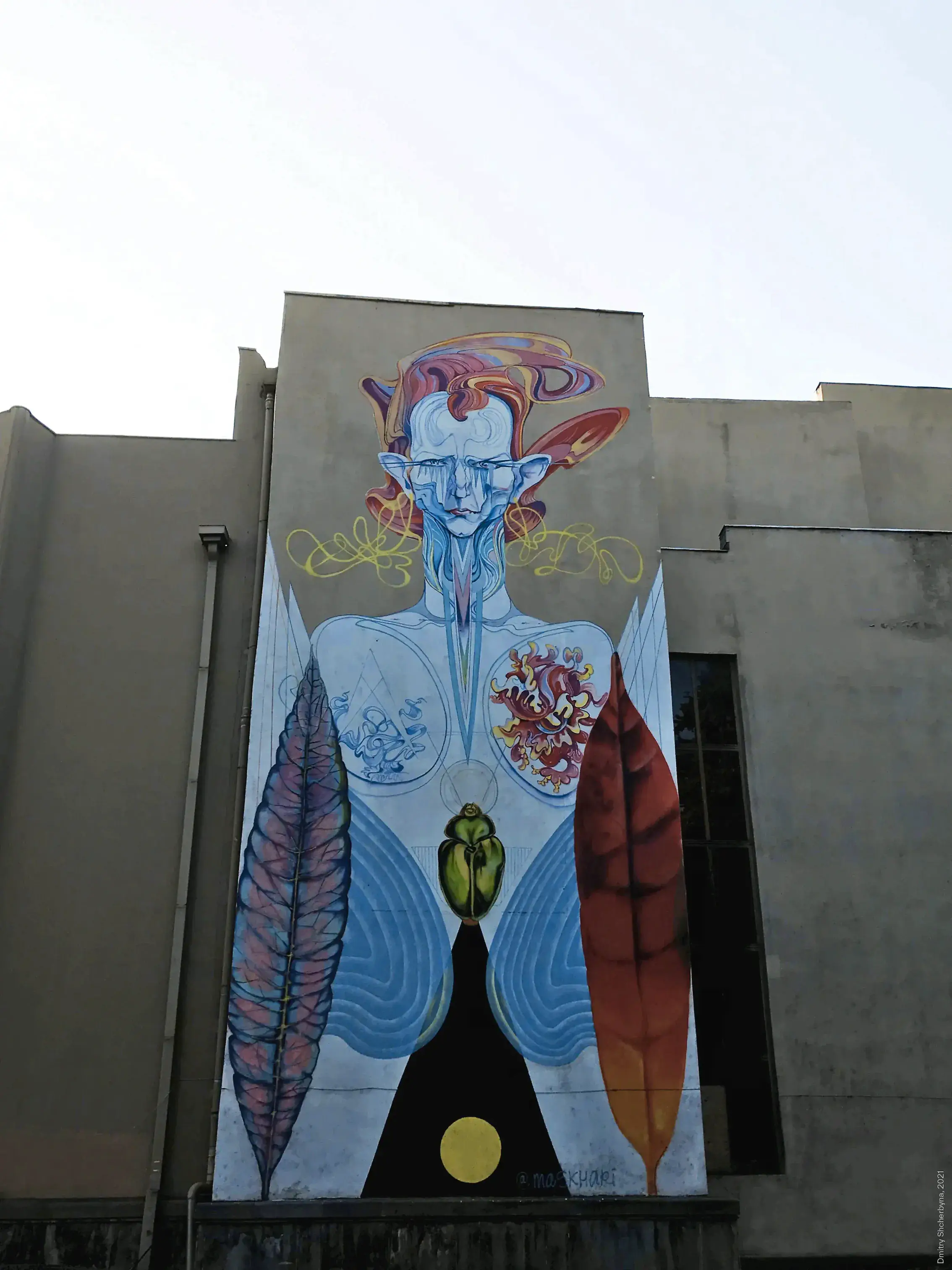 A tall surrealist mural featuring a pale blue humanoid figure with swirling multicolored hat, pointed ears, and elongated face.