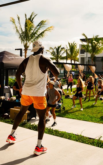 Fitness instructor leading an outdoor workout in South Florida, wearing bright orange shorts, with a group of participants exercising on a sunny day surrounded by palm trees.