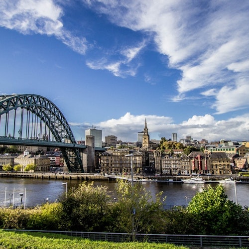 A bridge arches over a river with buildings, a church steeple, and trees on the far bank under a partly cloudy sky.