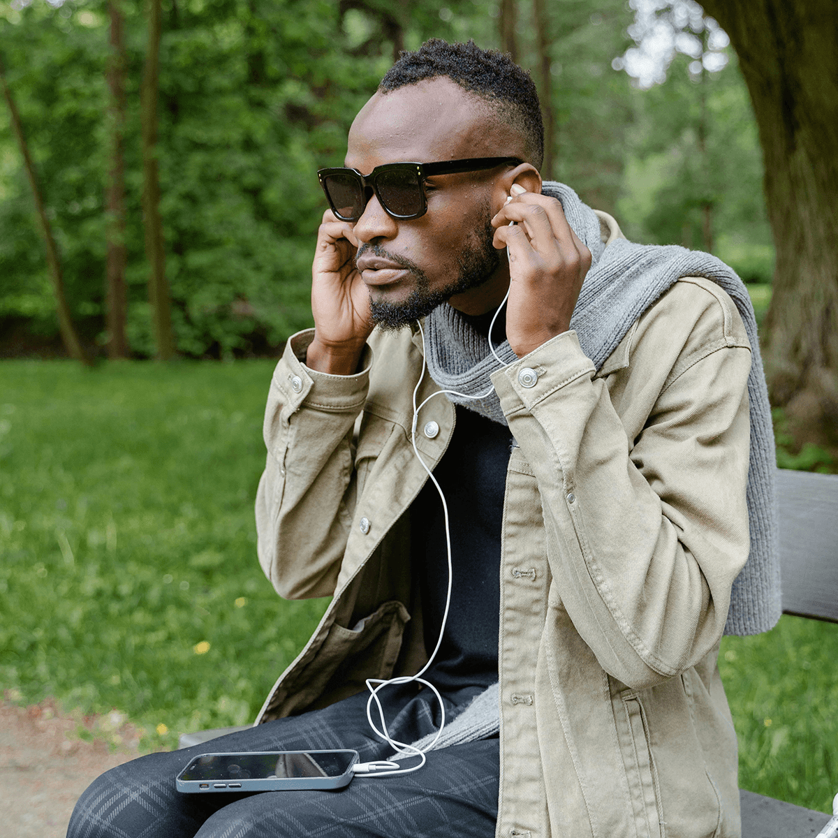 A visually impaired man sitting in a park, using a mobile phone with earphones plugged in