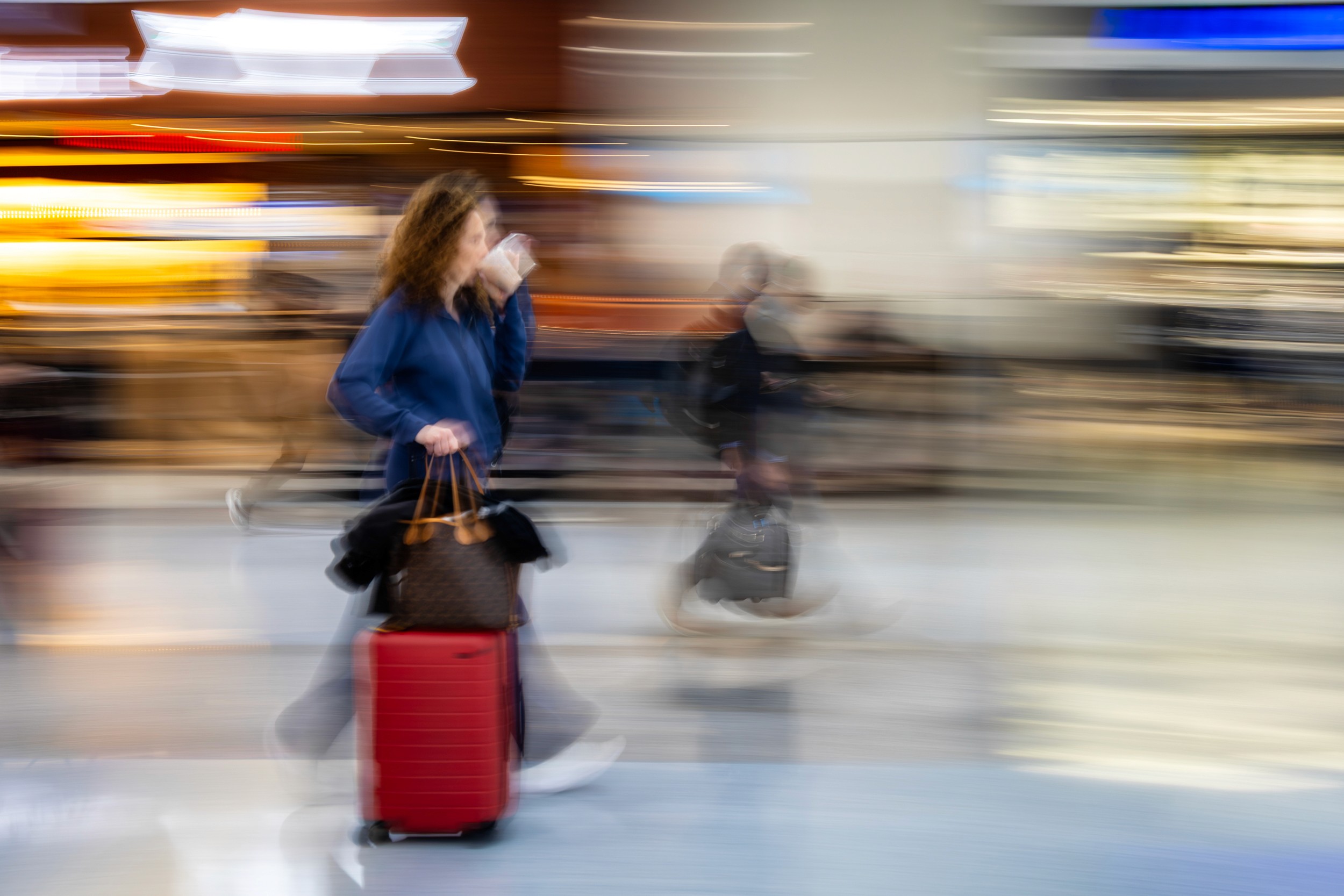 A traveler with a red suitcase stands still in a busy, blurred airport scene filled with people.