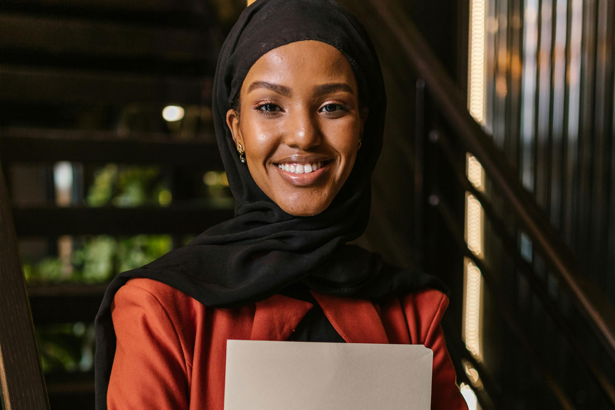 A smiling woman wearing a black hijab and red jacket, holding a laptop, in a warmly lit indoor setting.