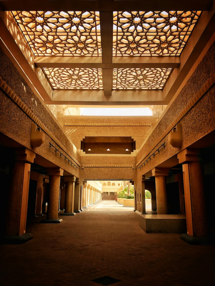 Sunlit courtyard corridor in Riyadh, Saudi Arabia, with geometric lattice skylights and warm sandstone columns.