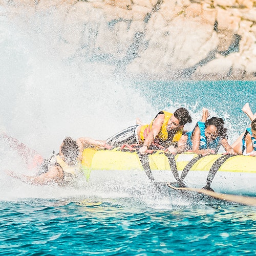 People in life jackets riding a yellow inflatable banana boat on water, with one person falling off, creating a splash. Rocky background.