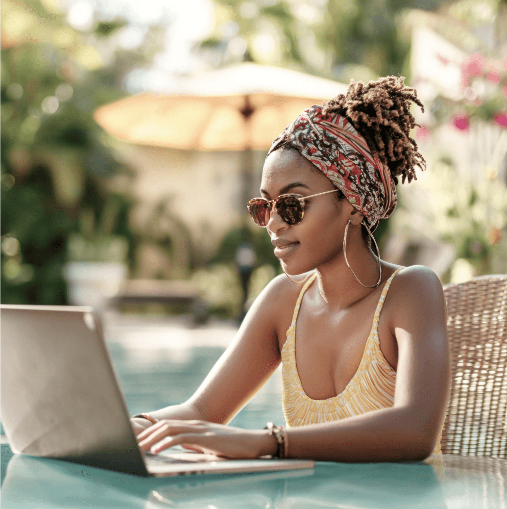A woman wearing sunglasses and a headscarf working on a laptop beside a swimming pool in a sunny, tropical setting.