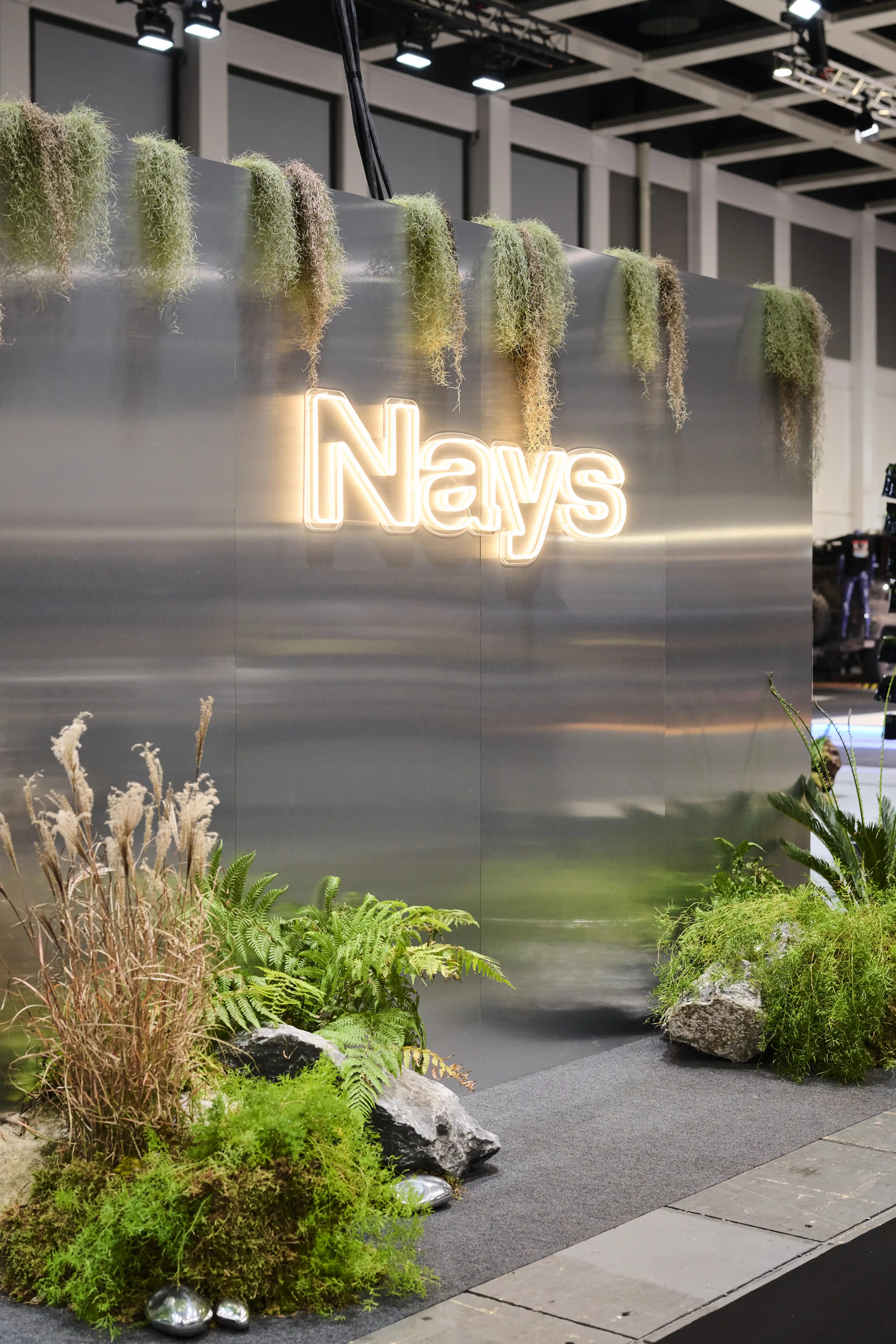 Brushed metal wall with glowing Nays logo, overhanging grasses and lush plant arrangements at the trade fair booth.