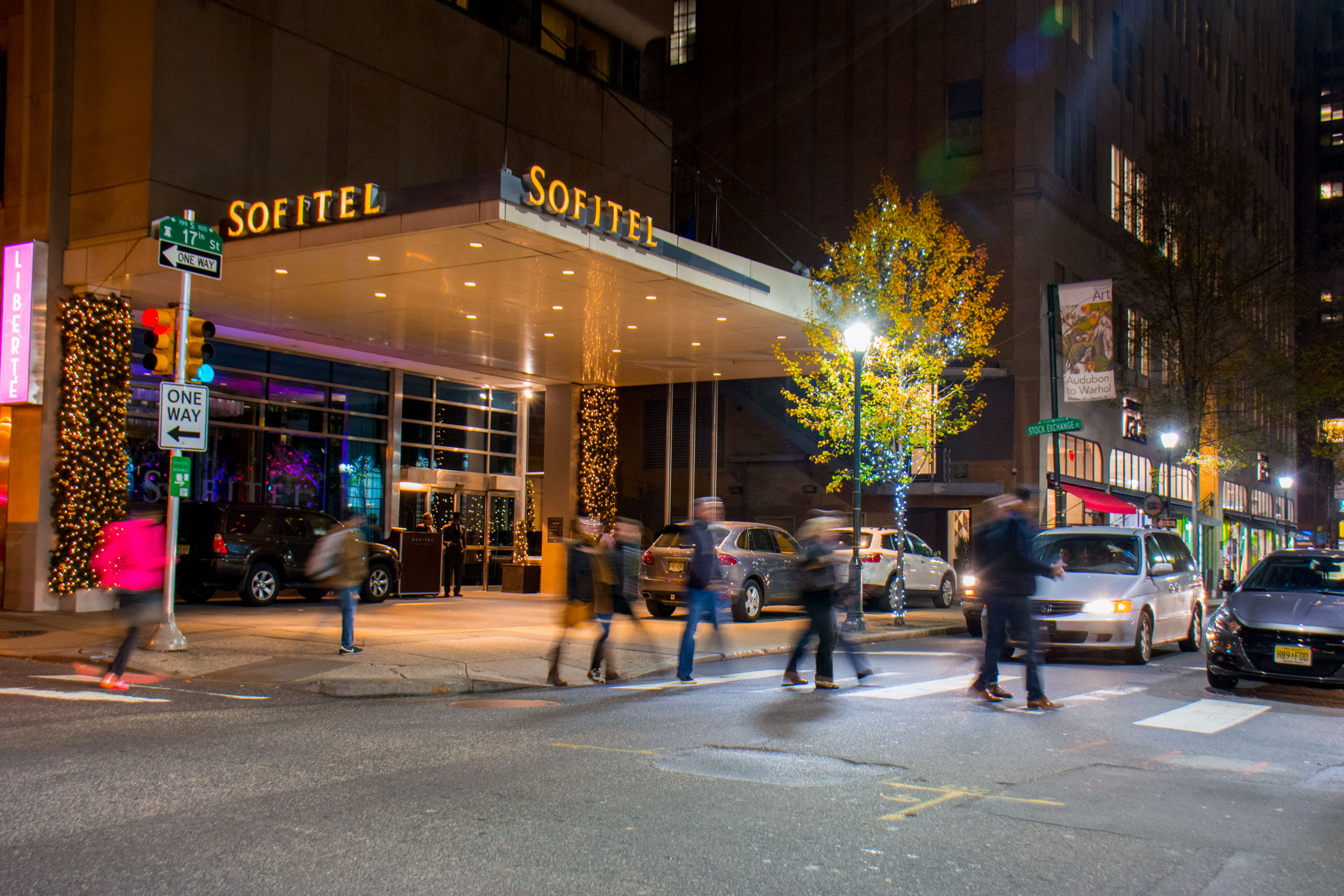 People walk along a city sidewalk at night in front of a Soffitel hotel with glowing signage and lit trees.