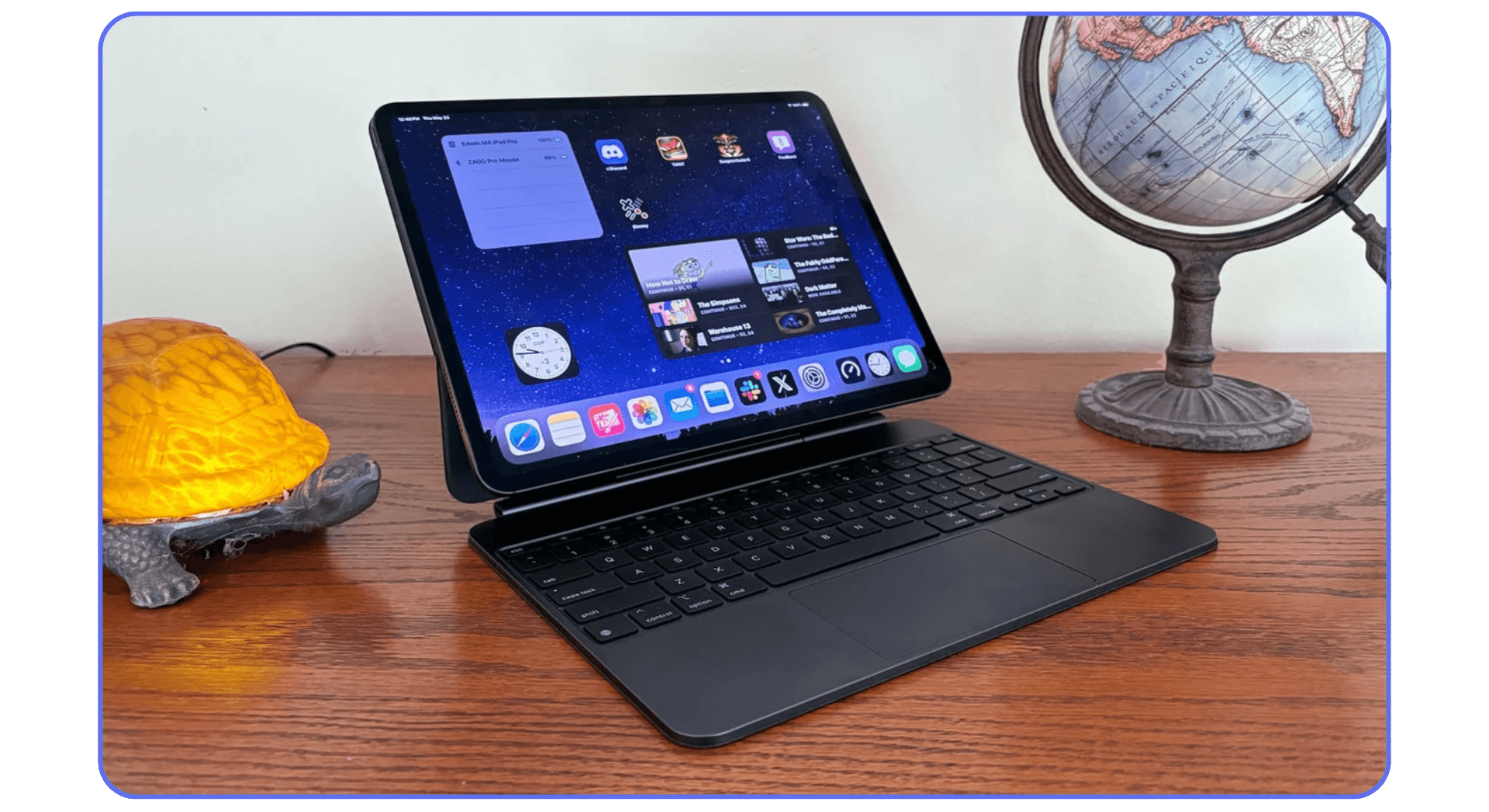 iPad connected to a Magic Keyboard on a wooden desk, displaying a home screen with widgets and apps. A globe and turtle-shaped lamp sit beside it.
