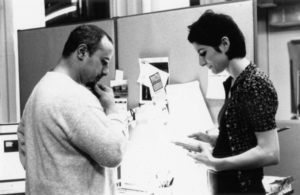 A black and white photo of a man and a woman checking some pinned notes on a wall.