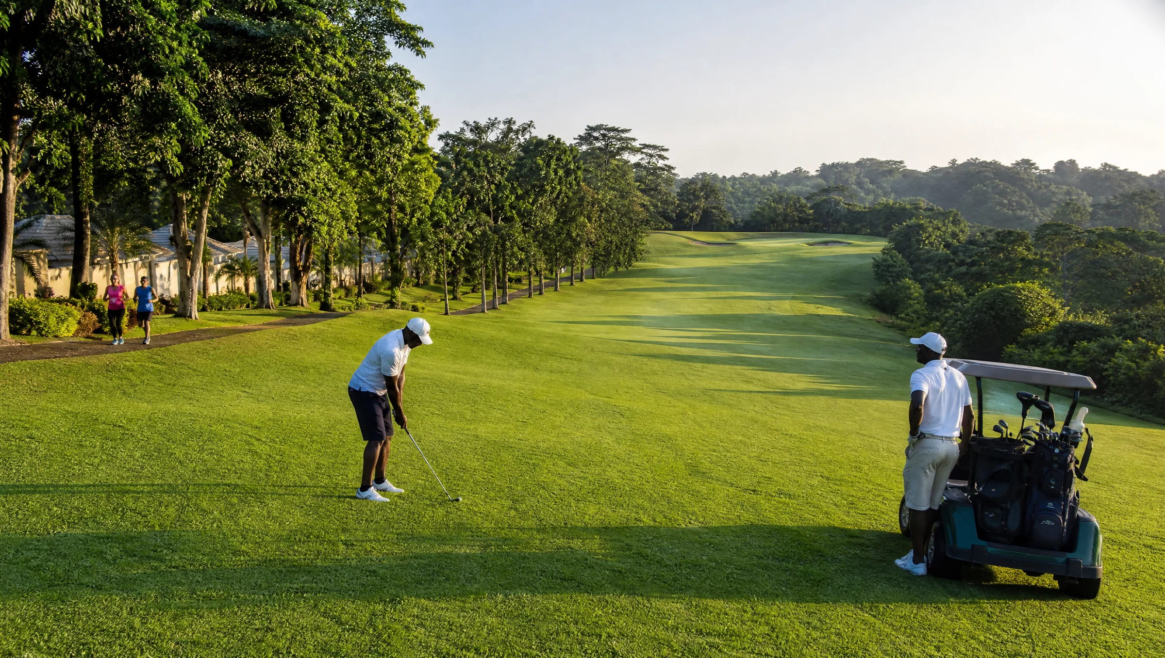 Golfers teeing off on a tree-lined fairway in early morning light, with joggers on a nearby path.