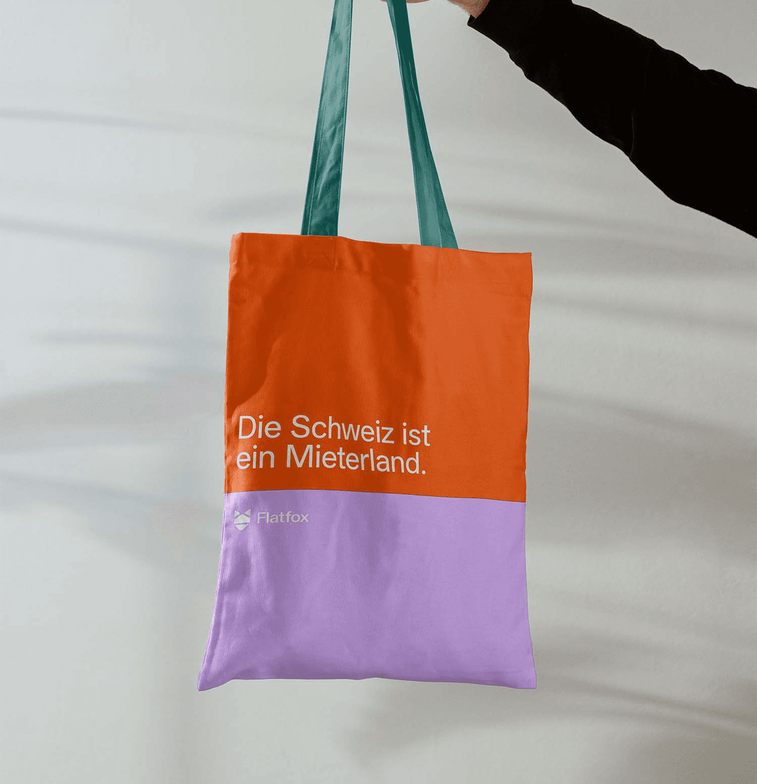 A hand holds a bicolor tote bag—orange on top and lavender on the bottom—featuring the German slogan "Die Schweiz ist ein Mieterland" (Switzerland is a land of tenants) and the Flatfox logo. This illustrates the studio's strategic copy and branding for the Swiss rental market.