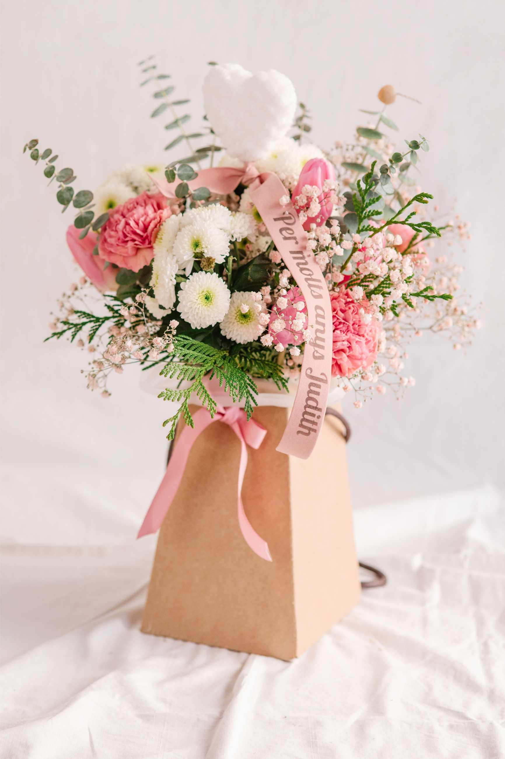 Composició floral delicada amb flors en tons suaus dins una bossa de cartró kraft, cinta rosa personalitzada amb missatge i cor blanc de feltre.