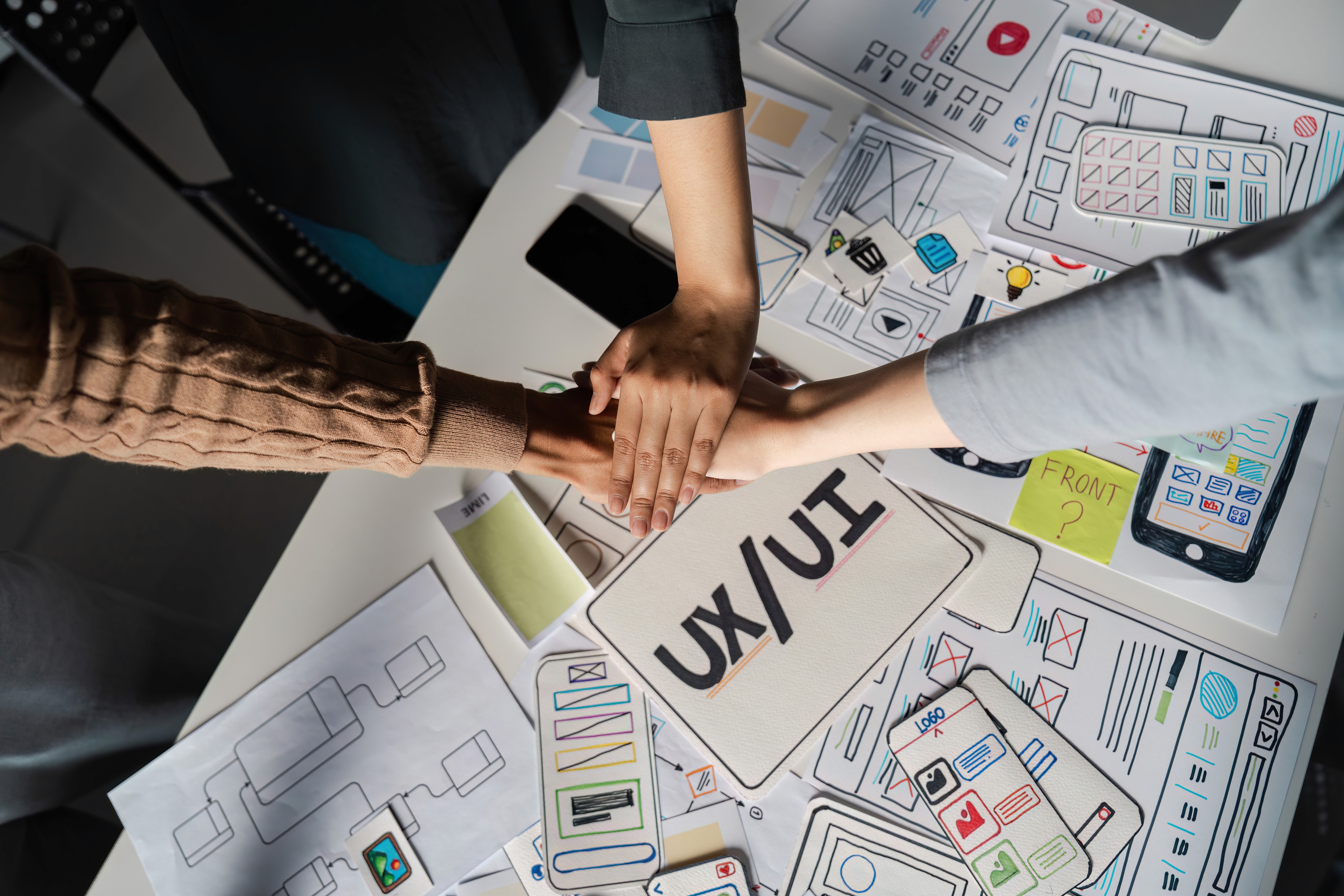 Team collaboration on UX/UI design with hands stacked in unity over mobile app wireframes and user interface mockups.