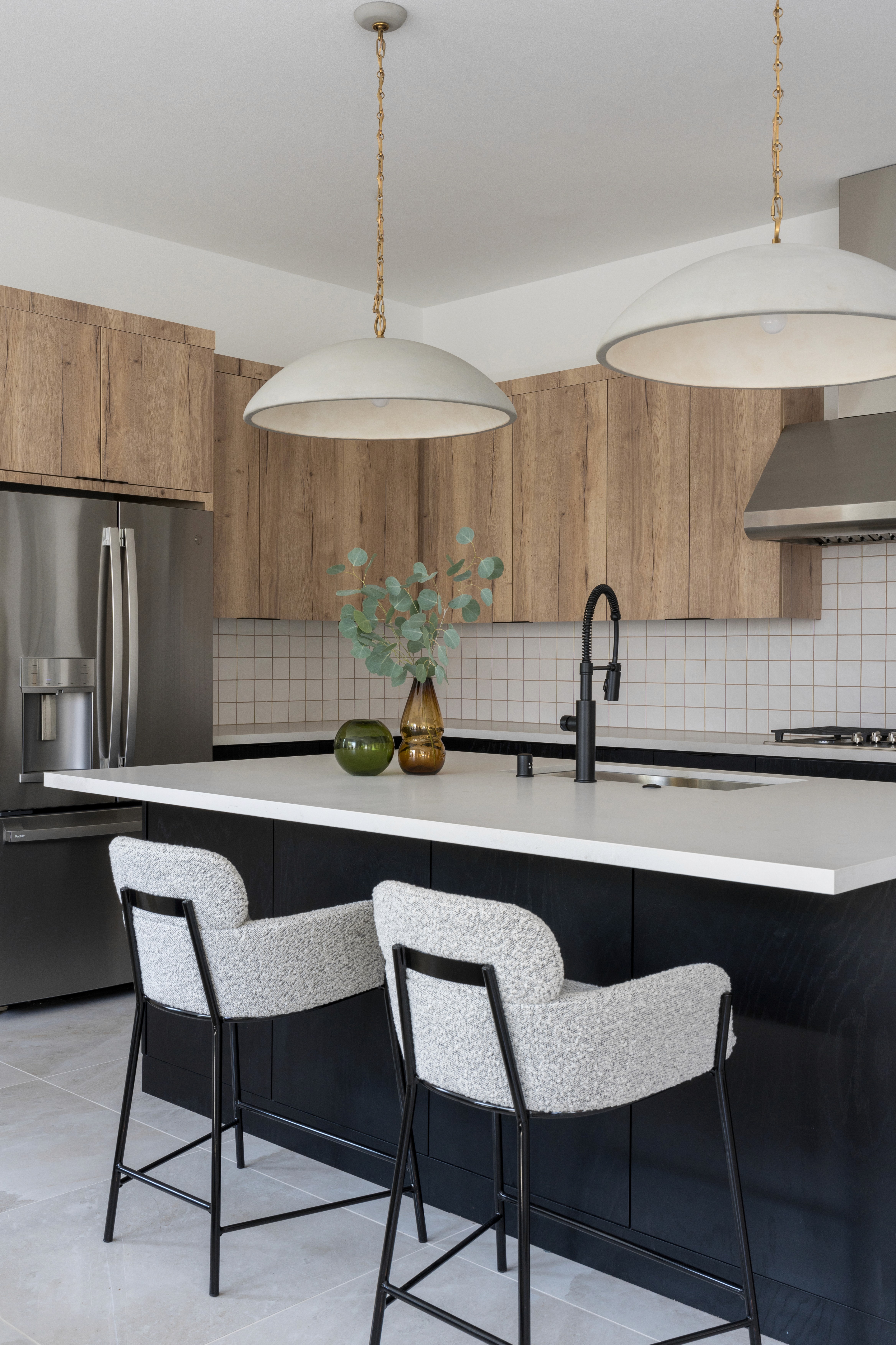 Kitchen with high neutral wood cabinets, white tile backsplash, low black wood cabinets, white countertops, grey boucle barstools, large pendant lighting with gold accents, and black brass faucet by Jubilee Interiors in Arcadia, California