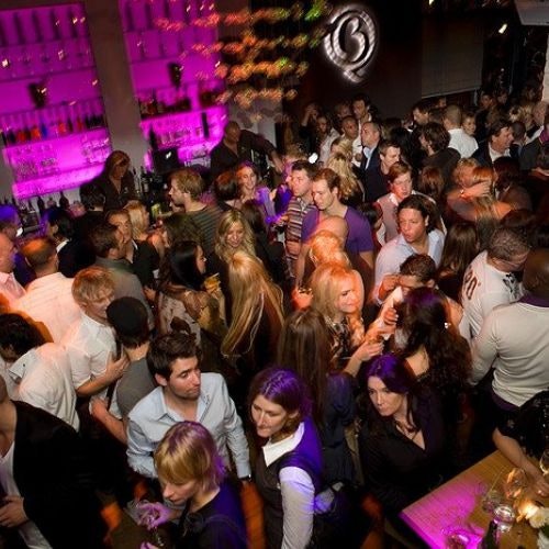 一个热闹的酒吧，人们在社交和饮酒；背景中，搁架上的瓶子被粉红色的灯光照亮。