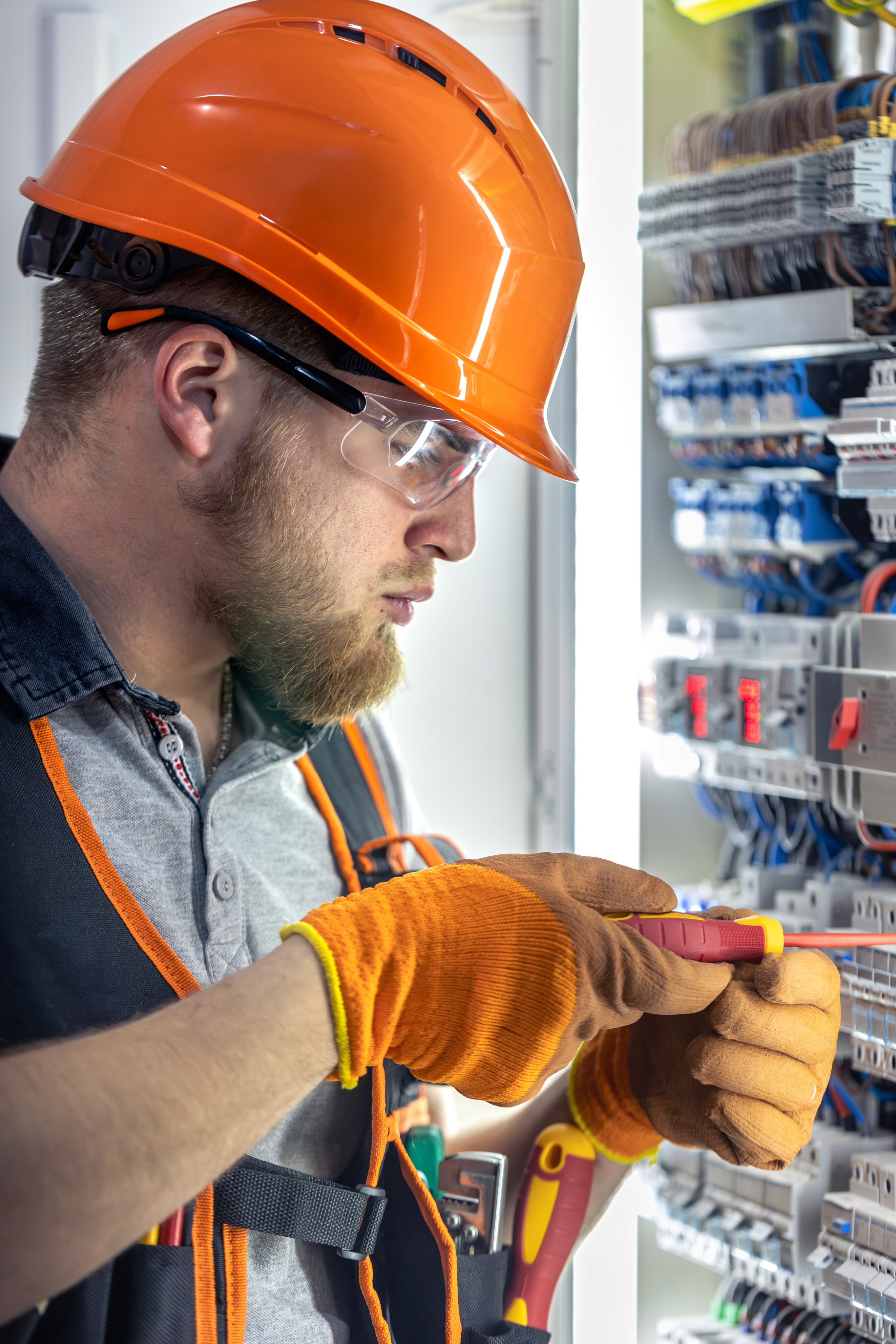 Electrician wearing an orange hard hat and gloves working on an open electrical panel
