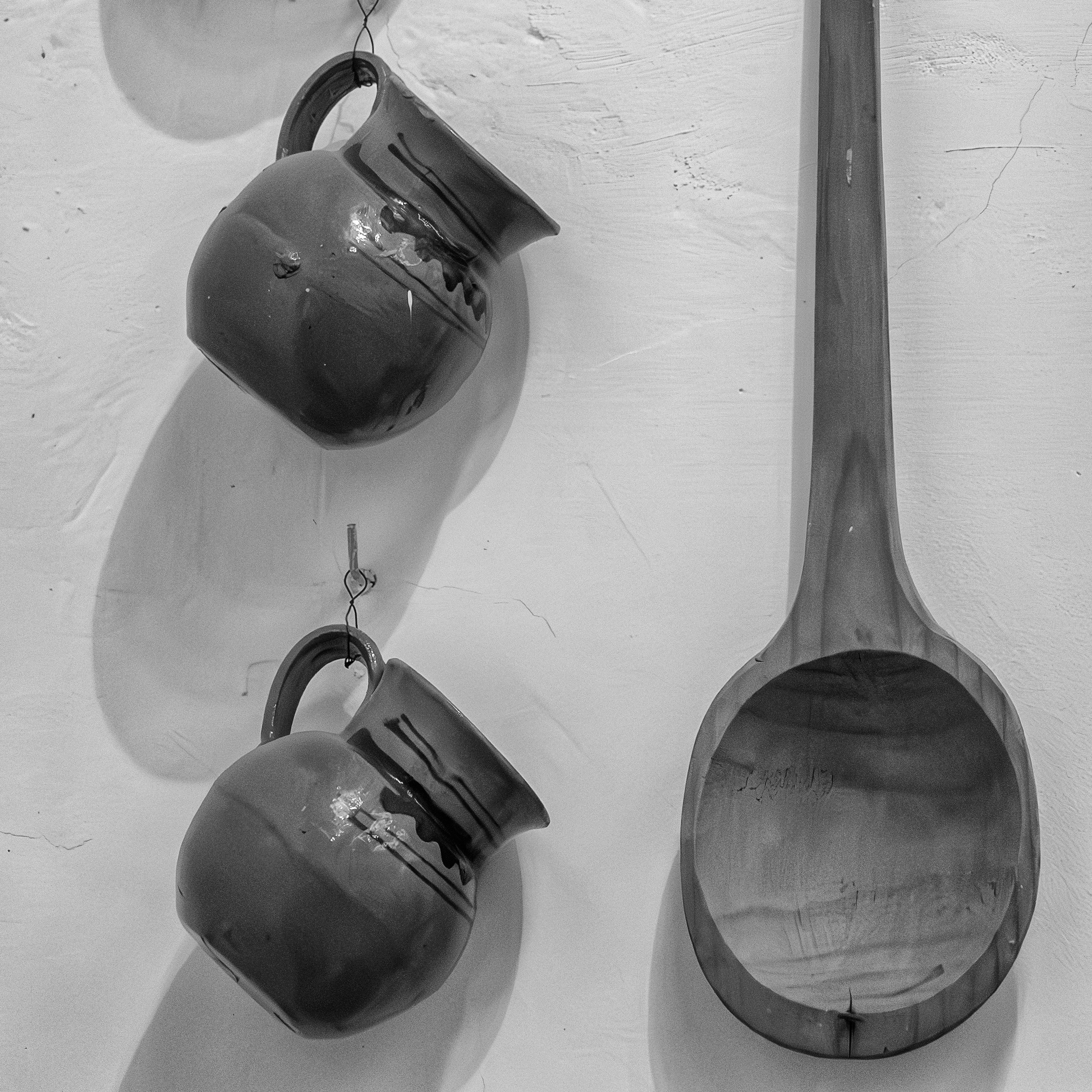 Black and white photo of two ceramic jugs and a large wooden ladle hanging on a textured wall, casting soft shadows.