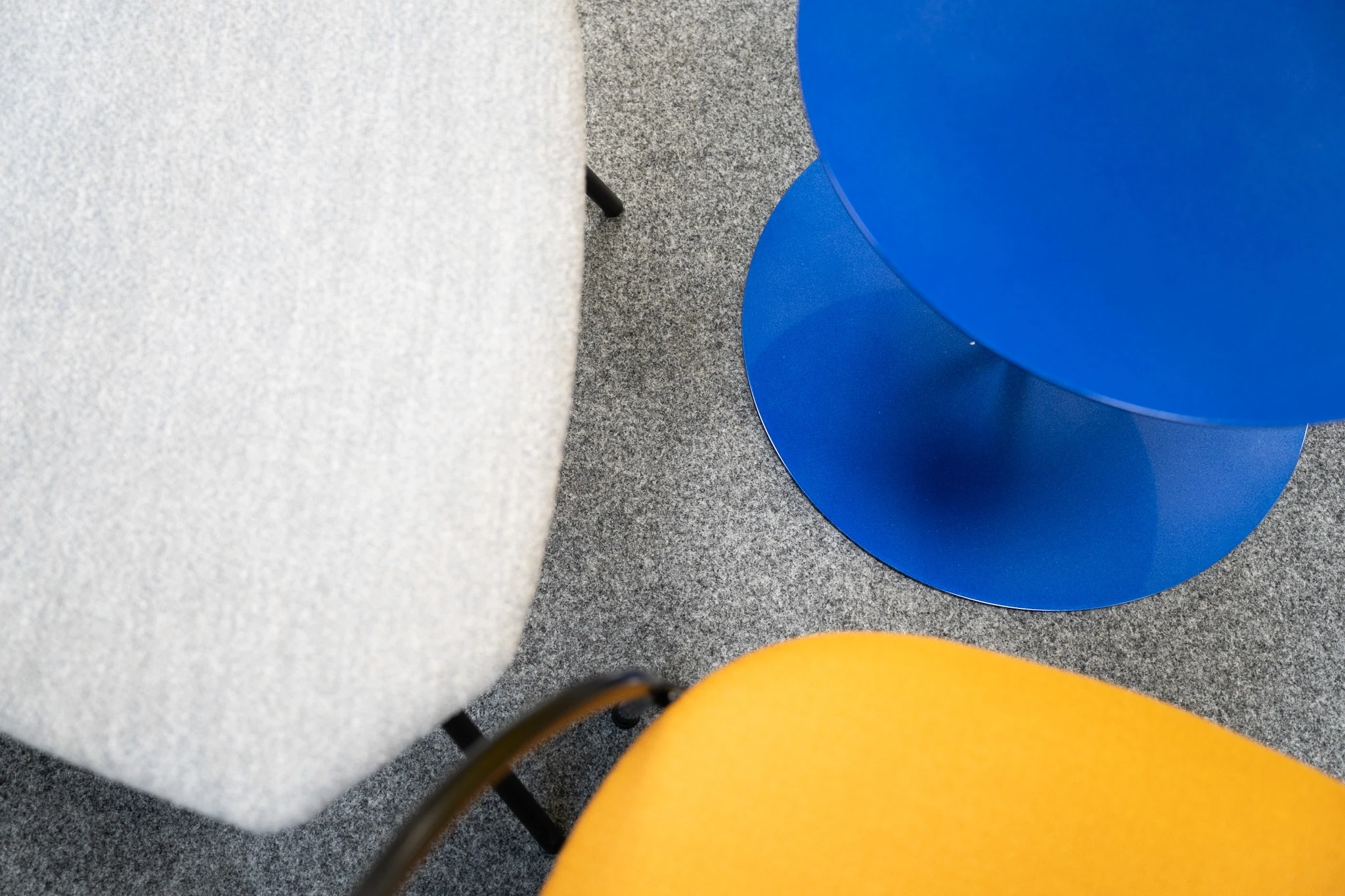 Top-down detail at NFON with a grey felt chair, a royal-blue metal side table and a mustard-yellow chair seat on speckled grey carpet.