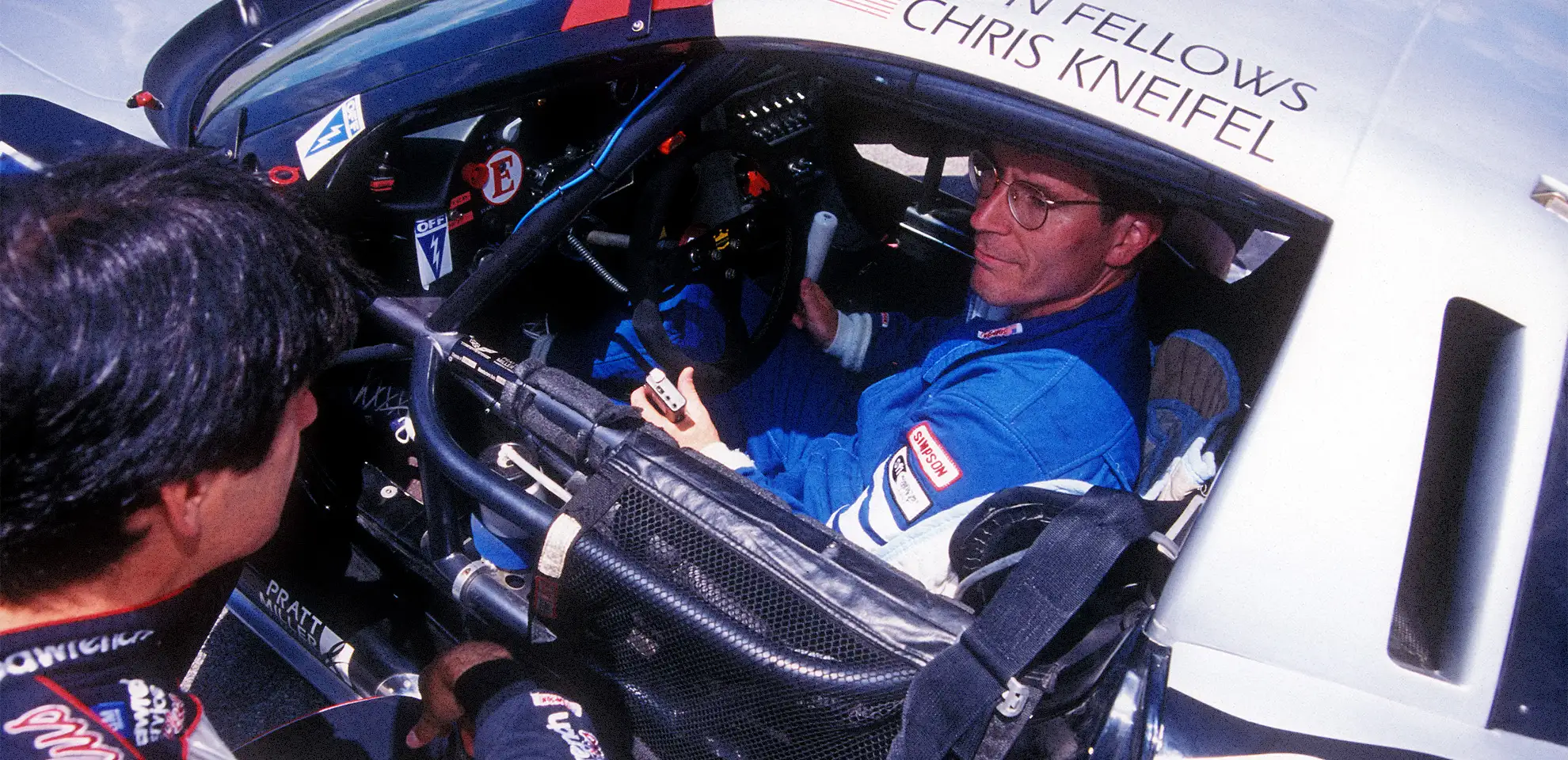 A race car driver in a blue suit sits inside a car, speaking with another person outside the vehicle.