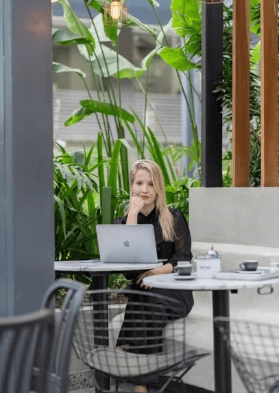 Person with blonde hair working on a laptop at an outdoor cafe table surrounded by tropical plants and modern lighting fixtures.