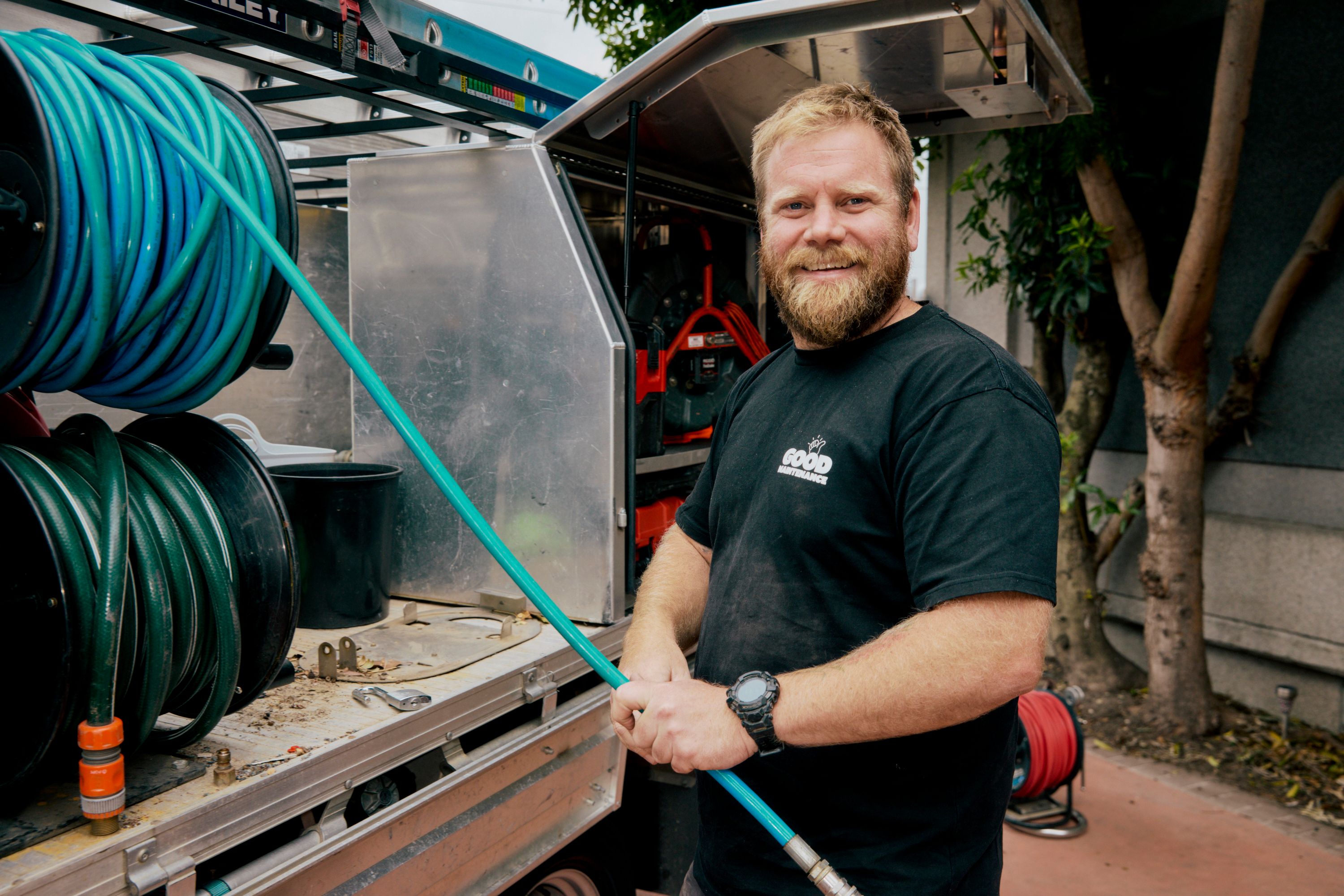 Portrait of a smiling Good Maintenance Technition, standing beside an open tray of tools, holding a hose.